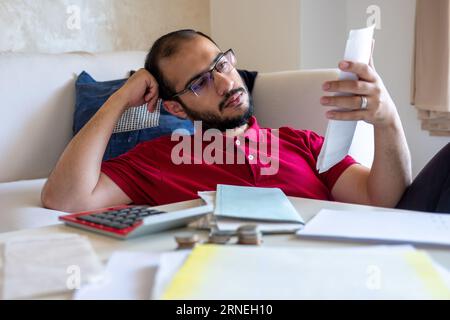 L'homme est frustré de ses factures de vie et de les calculer devant son canapé dans le salon Banque D'Images