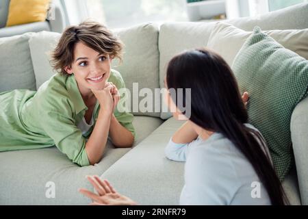 Photo de deux filles paisibles gaies assis couché canapé confortable communiquer raconter des histoires profiter de la maison de week-end à l'intérieur Banque D'Images