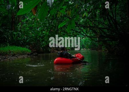 Homme inconnu dans un kayak rouge flottant vers une végétation luxuriante visible autour. Palmiers, mangroves, feuilles et autres plantes sous la pluie. Banque D'Images