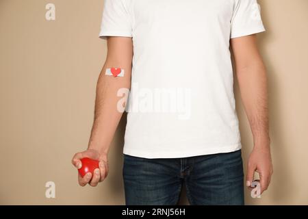 Concept de don de sang. Homme avec le plâtre adhésif sur le bras tenant le coeur rouge sur fond beige, gros plan Banque D'Images