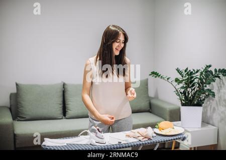 Une femme prend un petit déjeuner rapide tout en faisant des tâches ménagères. Une femme mange un savoureux hamburger tout en repassant des vêtements. Banque D'Images