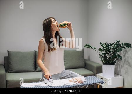 Une femme prend un petit déjeuner rapide tout en faisant des tâches ménagères. Une femme mange un savoureux hamburger tout en repassant des vêtements. Banque D'Images