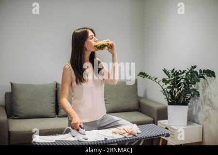 Une femme prend un petit déjeuner rapide tout en faisant des tâches ménagères. Une femme mange un savoureux hamburger tout en repassant des vêtements. Banque D'Images