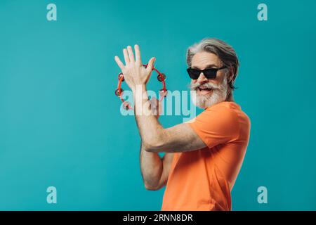 Un vieil homme barbu aux cheveux gris danse avec un tambourin. Photo de studio sur un fond coloré. Banque D'Images