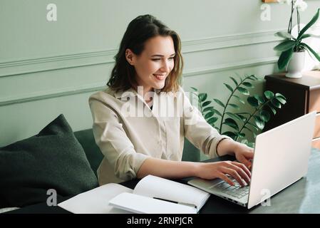 Jeune femme énergique multitâche. Saisie sur ordinateur portable et appels vidéo dans un cadre élégant en intérieur Banque D'Images