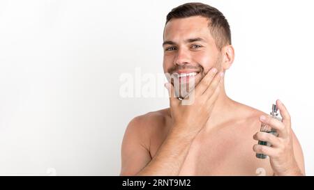 Homme heureux pulvérisant lotion après-rasage debout sur fond blanc Banque D'Images