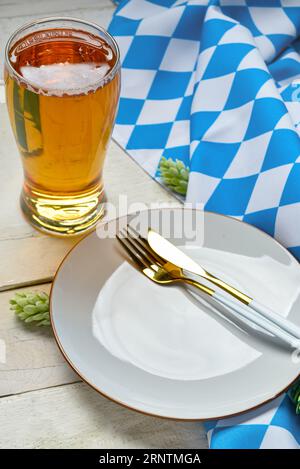 Assiette vide avec couverts, houblon, tasse de bière et nappe à carreaux bleus sur table en bois blanc Banque D'Images