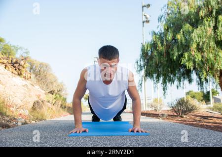 Un jeune homme caucasien fait des pompes sur son tapis dans un parc public. L'effort est visible sur son visage Banque D'Images