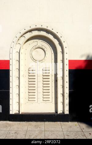 Une porte en métal blanc antique avec des rivets dans un cadre arqué, posée contre un mur peint avec une bande rouge et noire. Usine d'embouteillage de coca-Cola la Banque D'Images