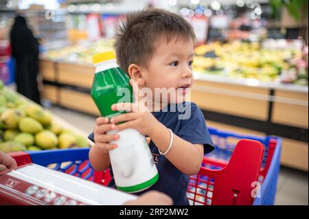 Enfant de deux ans adorable, souriant multiracial dans un t-shirt bleu assis dans un chariot avec des épiceries, tenant une bouteille avec de petites mains, Banque D'Images