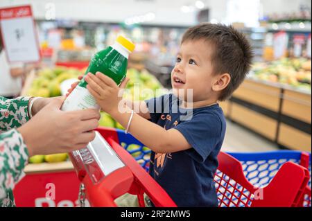 Enfant de deux ans adorable, souriant multiracial dans un t-shirt bleu assis dans un chariot avec des épiceries, tenant une bouteille avec de petites mains, Banque D'Images