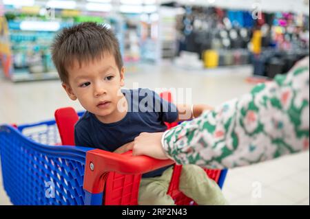 Deux ans adorable enfant multiracial dans un t-shirt bleu assis dans un chariot avec épicerie, dans l'épicerie Banque D'Images