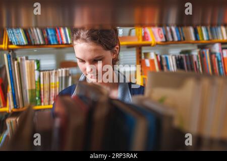 Écolière cherchant et choisissant des livres dans une étagère dans la bibliothèque. Éducation, littérature et nouveaux concepts d'édition. Banque D'Images