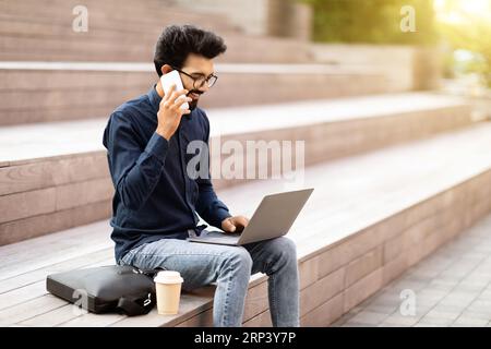 Souriant jeune homme indien Manager avec des appels d'ordinateur portable par téléphone Banque D'Images