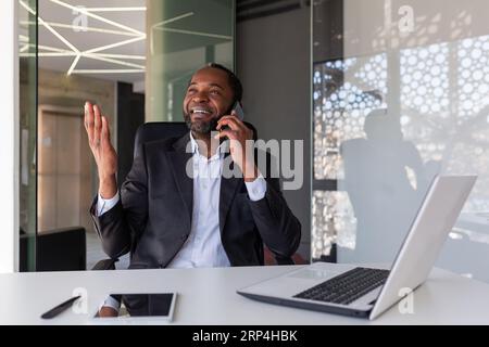 Patron expérimenté mature parlant au téléphone au lieu de travail à l'intérieur du bureau, homme d'affaires souriant ayant une conversation avec des partenaires et des collègues, satisfait des résultats de la négociation. Banque D'Images