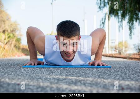 Un jeune homme caucasien fait des pompes sur son tapis de sport dans un parc public. Il sourit à la caméra Banque D'Images