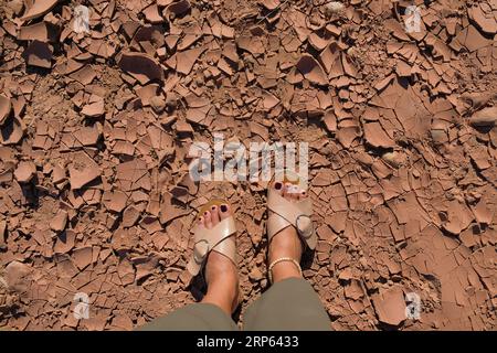 Les pieds féminins avec des sandales se tiennent sur un sol craquelé et desséché à ait Ben Haddou, un village d'argile historique au Maroc. Concept de réchauffement climatique. La personne marche o Banque D'Images