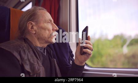 Homme âgé voyageant en train et prenant des photos du paysage par la fenêtre à l'aide d'un smartphone Banque D'Images