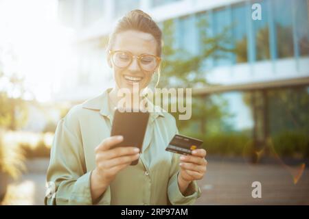 souriant femme moderne travailleur dans le quartier des affaires en blouse verte et lunettes avec carte de crédit à l'aide d'un smartphone. Banque D'Images