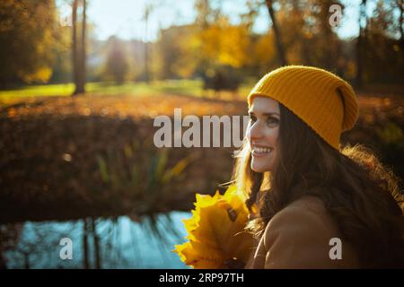 Bonjour automne. femme moderne souriante en manteau beige et chapeau orange avec des feuilles jaunes d'automne assis à l'extérieur sur le parc de la ville en automne. Banque D'Images