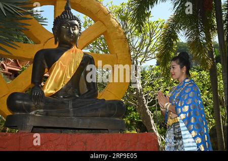 Jolie femme asiatique en costume national thaïlandais debout pose et rend hommage devant la statue de Bouddha peinte en noir à l'extérieur. Banque D'Images