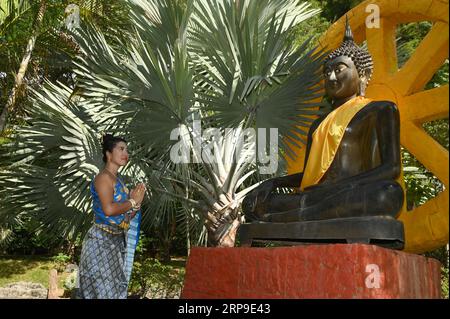 Jolie femme asiatique en costume national thaïlandais debout pose et rend hommage devant la statue de Bouddha peinte en noir à l'extérieur. Banque D'Images