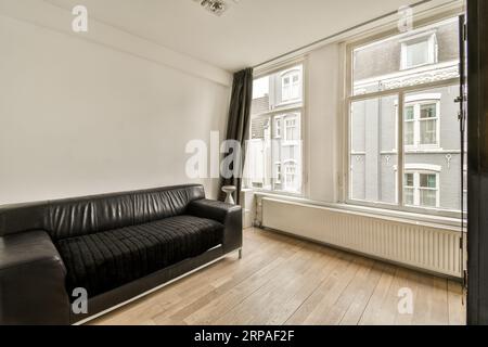 un salon avec parquet et murs blancs, il y a un canapé en cuir noir devant la fenêtre Banque D'Images