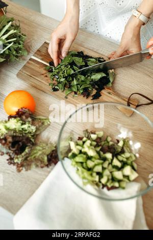 Une femme coupe le basilic et les herbes avec un couteau sur une planche à découper, à côté de légumes et d'une assiette de légumes hachés. Banque D'Images