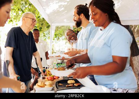 Les personnes volontaires qui aident les personnes défavorisées en fournissant de la nourriture et des aliments à ceux qui en ont besoin. Les bénévoles distribuent de la nourriture avec des sourires, assurant l’inclusion des personnes handicapées et des sans-abri. Banque D'Images