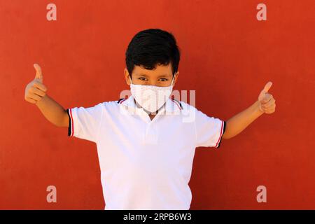 Joyeux et pensif Latino garçon de 8 ans portant une chemise blanche d'uniforme scolaire et un masque facial pour la protection contre la pandémie de Covid-19 à la rentrée scolaire Banque D'Images