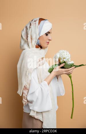 vue latérale de femme musulmane à la mode en foulard et blouse blanche tenant fleur d'hortensia sur beige Banque D'Images