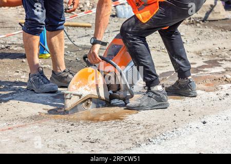 Un coupeur circulaire à essence entre les mains d'un ouvrier coupe la surface de la route en asphalte c oncrete par une journée ensoleillée d'été. Espace de copie. Banque D'Images