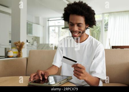 Heureux jeune homme afro-américain regardant la carte de crédit dans sa main tout en étant assis sur le canapé à la maison et en entrant le numéro sur le site Web Banque D'Images