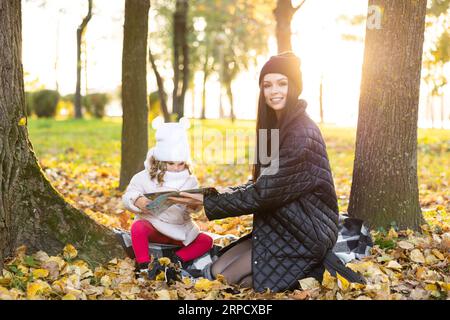 Famille heureuse s'amusant dans le parc d'automne. Une mère lit un livre avec sa fille mignonne assise sur la pelouse dans le parc Banque D'Images