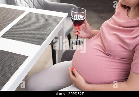 Une femme enceinte s'assoit dans la cuisine à la table et boit de l'alcool. Mauvaise habitude pendant la grossesse. Banque D'Images