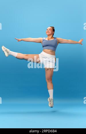 Photo pleine longueur de souriante femme en surpoids portant sportswear saut et faisant des exercices isolés sur fond bleu studio. Banque D'Images
