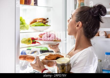 Le vendredi soir, une femme très affamée en pyjama choisit des collations, des steaks, des beignets et une boîte de cornichons dans le réfrigérateur. Banque D'Images