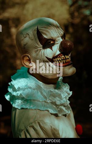 un clown maléfique chauve, vêtu d'un costume gris avec un froissé blanc et des pompons rouges, se retourne pour fixer l'observateur avec un sourire effrayant, dans le w Banque D'Images