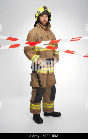 Pompier sérieux en train de se tenir derrière le ruban de signalisation rouge et blanc. Vue avant du sauveteur barbu regardant la caméra depuis la zone fermée par le ruban de mise en garde, sur fond blanc. Concept de lieu de travail. Banque D'Images