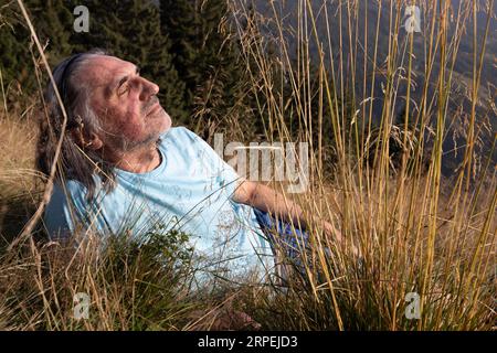 Homme adulte aux cheveux gris avec de longs cheveux reposant sur un champ avec de l'herbe haute dans la lumière du soleil du soir Banque D'Images