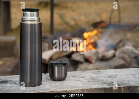 Bouteille thermos en acier rayée sur un banc en bois à côté du feu de camp Banque D'Images