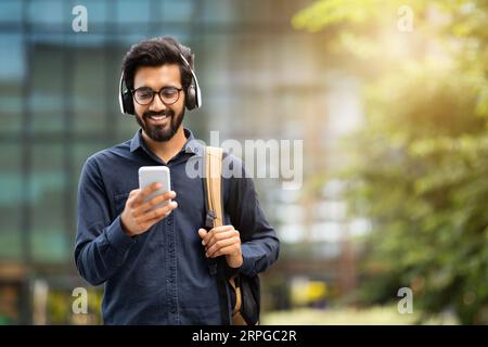 Souriant jeune homme indien portant des écouteurs tenant le téléphone tout en marchant Banque D'Images