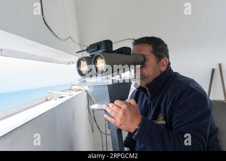 Un belvédère employé par des compagnies d'observation des baleines regarde à travers des jumelles alors qu'il regarde l'océan Atlantique pour des observations de baleines près d'Agua de Pau, île de Sao Miguel, Açores, Portugal. Les postes d'observation de la chasse à la baleine étaient autrefois utilisés par l'industrie baleinière pour chasser les baleines et font maintenant partie de l'éco-industrie d'observation des baleines, qui a connu un succès et est durable. Banque D'Images