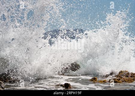 Une vague se brise sur les grosses pierres dans l'eau, des milliers de gouttes d'eau scintillent dans la lumière du soleil, l'île silhouette dans l'arrière-plan, snapshot - Région Banque D'Images