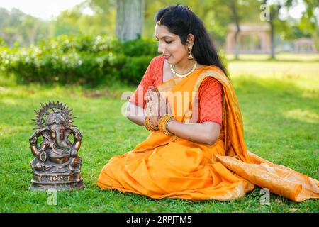 Heureuse jeune femme indienne portant Saree assise adorant lord Ganesha idole en plein air au parc célébrant le festival Ganesh Chaturthi. Banque D'Images