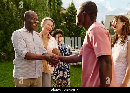 Deux hommes afro-américains heureux se serrant la main tout en se tenant devant la caméra contre leurs épouses et garçon mignon et saluant l'un l'autre Banque D'Images