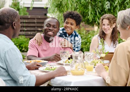 Jeune couple interculturel et leur fils mignon en vêtements décontractés regardant l'homme mûr et la femme assis devant eux par la table servie Banque D'Images