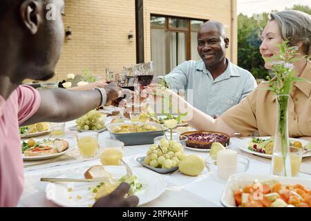 Heureux couple mature avec des verres à vin griller avec leur fils et sa femme assis à table servi avec de la nourriture maison pendant le dîner en plein air Banque D'Images
