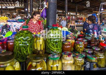 Un commerçant de marché vêtu d’un chemisier rouge à motifs floraux se tient derrière des contenants en verre et des bocaux de légumes marinés sur le marché couvert des producteurs de l’avenue Chavchavadze à Telavi, en Géorgie. Les bocaux contiennent de longues gousses vertes, des cornichons, des piments forts et une pâte rouge. Le commerçant regarde une femme en robe fleurie devant le stand à droite. En arrière-plan, une jeune vendeuse en noir et un homme en chemise à carreaux se tiennent à un stand de chou blanc. Des Churchkhela pendent sous le toit. Pour fabriquer ce dessert traditionnel géorgien, des noix ou des noisettes sont enfilées sur une ficelle. Les brins sont Banque D'Images