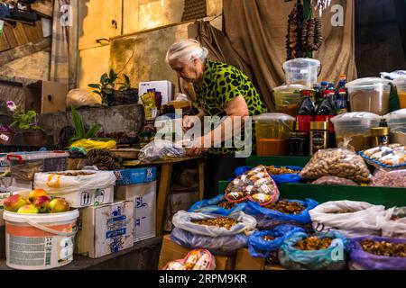 Un marchand de marché âgé aux cheveux blancs et portant un chemisier à motifs verts et noirs est penché sur une table en bois à son étal sur le marché couvert des producteurs de l'avenue Chavchavadze à Telavi, en Géorgie. De sa main droite, il attrape de petits produits emballés. Des noix et des fruits secs sont posés dans des sacs ouverts au premier plan, avec un sac en forme de cœur contenant des bonbons. À droite se trouvent des bacs en plastique contenant des produits orange et bruns, des bouteilles de cola et des bocaux de café instantané. Des anneaux de tranches séchées enfilées pendent en haut. Des pommes dans un seau et des plantes en pot se trouvent à gauche. Un toit incurvé recouvre le mai Banque D'Images